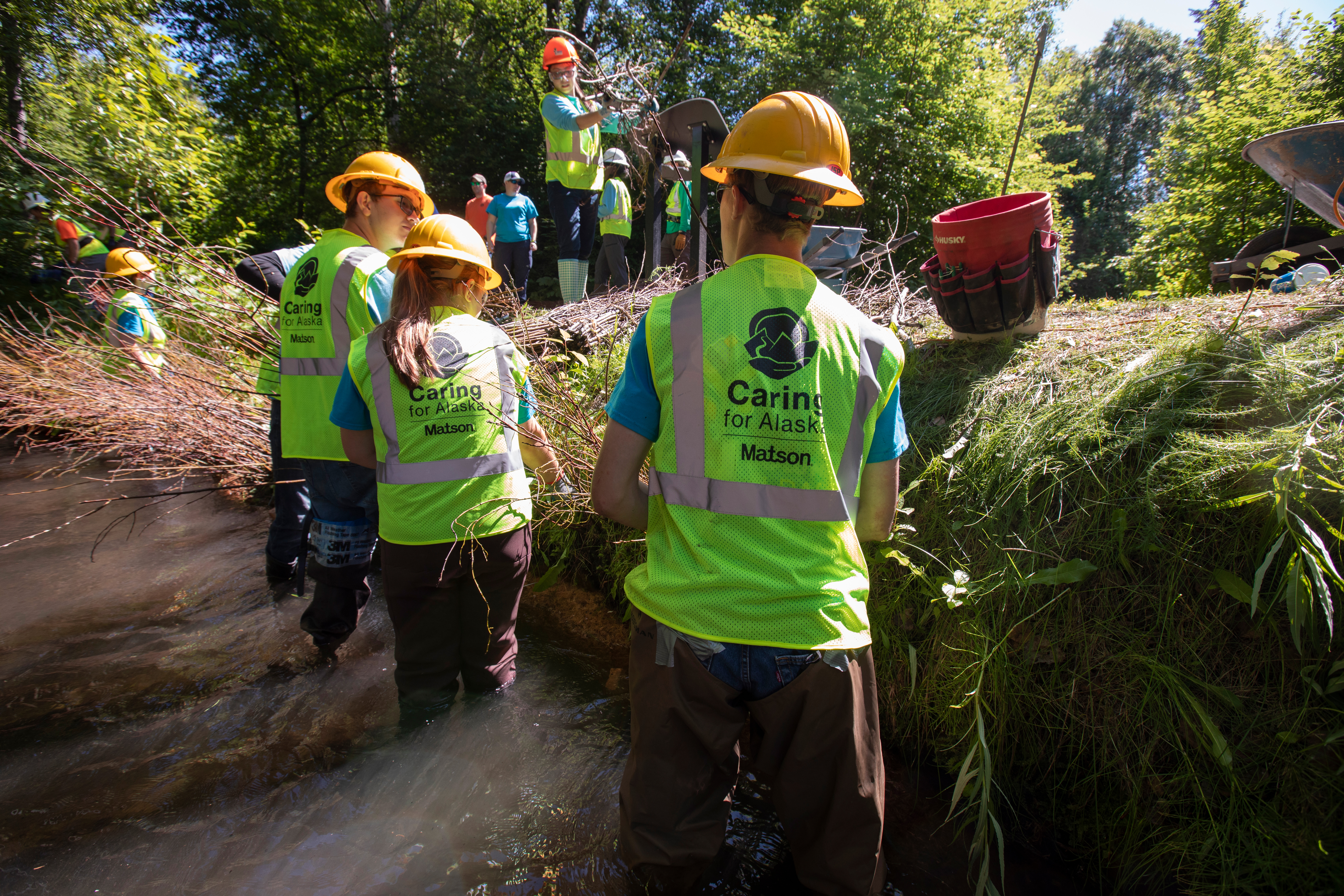 Youth Employment in Parks participants wear green safety vests and yellow hard hats while standing in Chester Creek to remove invasive species and conduct habitat restoration.